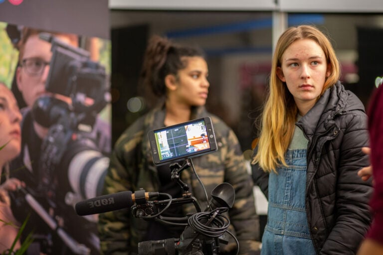 Alt text: A young woman with a focused expression stands in the foreground at a video production workshop. She is wearing a black jacket over a denim overall dress. Behind her, a professional video camera on a tripod is in focus, with its screen displaying a colorful video feed. To the left, a backdrop features a life-size photograph of a videographer holding a camera, creating a layered effect. Another participant is partially visible in the background, observing the environment. The setting suggests an educational or behind-the-scenes look at video production.
